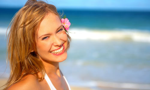 Close-up picture of a beautiful woman, happy with her face lift with neck lift in San Jose, Costa Rica.  The woman has long sandy blonde hair and is looking directly at the camera with the ocean behind her.