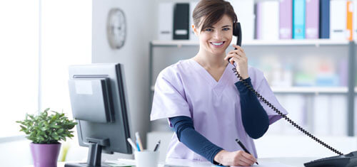Picture of a receptionist representing Top Plastic Surgeons in San José, Costa Rica.  The woman has short brown hair, is wearing a hospital smock and is standing at the receptionist desk while smiling at the camera.